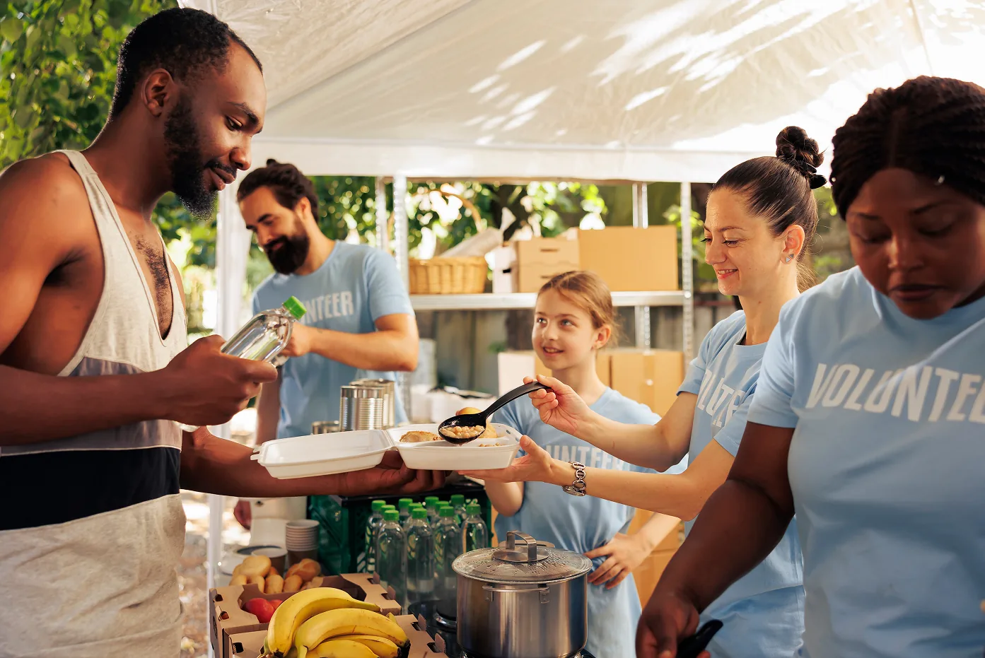 Volunteers serve fresh food under a canopy at a sunny community meal event.