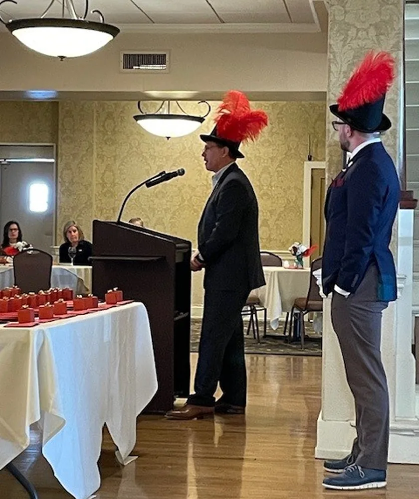 Formal event with two men wearing tall black hats with red plumes.