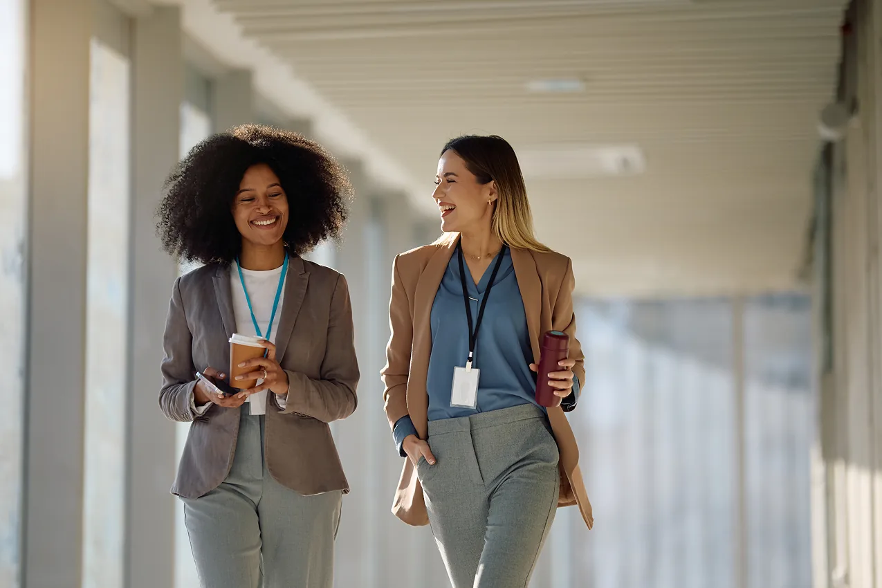 Two joyful colleagues walk together through a bright, modern office hallway.