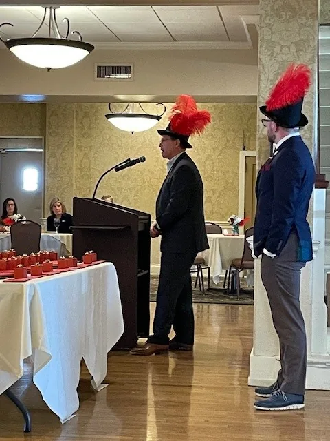 Man in red feathered hat speaks at podium during formal ceremony.